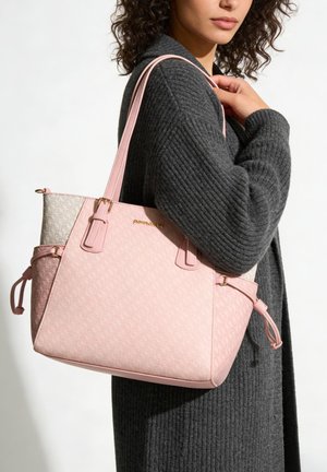 Frau trägt einen dunkelgrauen Strickpullover und hält eine große pink-beige gemusterte Tragetasche mit goldenen Beschlägen auf der Schulter.