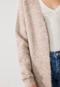 Cardigan en moumoute rose clair avec des manches côtelées et un devant ouvert, superposé sur un haut blanc à col en v côtelé, associé à un jean en denim bleu.
