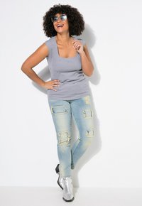 Haut sans manches gris avec des détails en perles, associé à un jean léger en denim déchiré avec un patchwork. Le mannequin porte des bottines argentées et des lunettes de soleil.