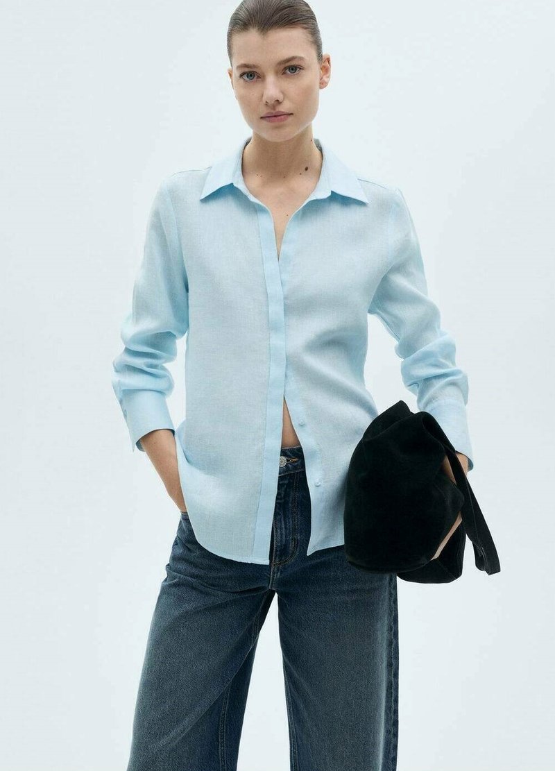 Camicia button-up blu chiaro in un tessuto morbido, con colletto e maniche lunghe. Abbinata a jeans in denim scuri e una borsa nera.