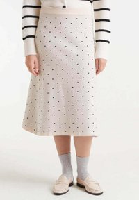 Falda crema hasta la rodilla con lunares negros, hecha de material suave. Combinada con calcetas grises y mocasines de color claro.