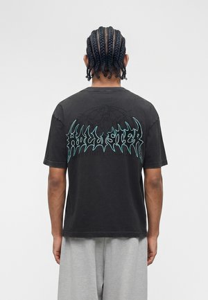 Osoba s ispletenom kosom nosi crnu majicu s plavim apstraktnim tekstom na leđima i svijetlo sive hlače, stoji na jednobojnoj pozadini.