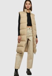 Beige puffer vest met hoge kraag, gestikt ontwerp en een lange snit. Gekoppeld aan beige wijde broek en zwarte plateau laarzen.