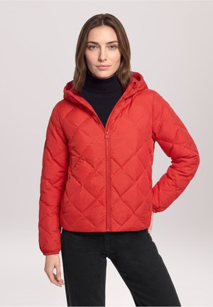 Frau mit braunen Haaren, die eine rote gesteppte Jacke mit Kapuze und schwarze Hose trägt, steht mit einer Hand in der Hüfte vor hellem Hintergrund.
