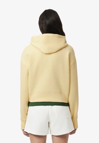 Gekürzte gelbe Kapuzenjacke aus weichem Material mit einer großen Kapuze und einem grünen Saum, getragen über weißen Shorts. Die Rückansicht zeigt das Logodesign.