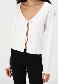 Vit kort cardigan, ribbad textur, djup V-hals, knytning vid bysten, vågkantad nederkant och ärmslut, kombinerad med svarta leggings.