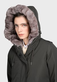Ciemnozielona parka z kapturem wyłożonym sztucznym futrem w jasnoszarym kolorze. Posiada zamek błyskawiczny i zapięcie na guziki, regulowane sznurki oraz pikowaną teksturę.