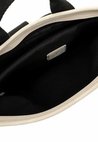 Juodas audinio maišelis su smėlio spalvos apvadu, turintis lygų vidų ir mažą identifikavimo etiketę. Paprastas ir funkcionalus dizainas.