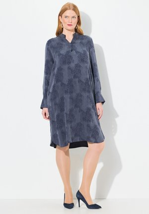 Langarmkleid in Dunkelblau mit einem dezenten Blumenmuster, ausgestattet mit einem Stehkragen und einem leicht ausgestellten Schnitt. Kombiniert mit blauen High Heels.