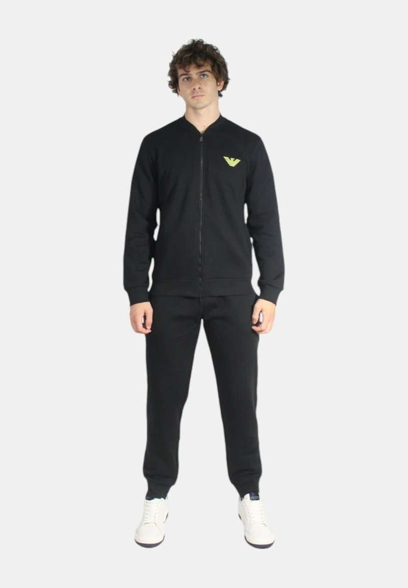 Felpa nera con zip, polsini a coste, pantaloni coordinati e sneakers bianche. Presenta un logo verde sul petto. Texture del tessuto liscia.