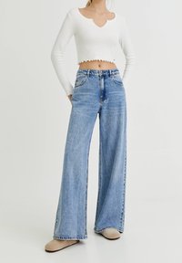 Top blanco acortado con un dobladillo en forma de ondas, combinado con jeans de mezclilla azul de pierna ancha y cintura alta. Unos zapatos de slip-on en color marrón claro completan el atuendo.