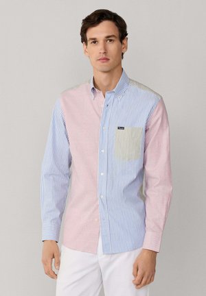 Homme portant une chemise à manches longues à boutons avec des rayures verticales bleues, roses et beiges, associée à un pantalon blanc.