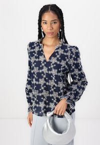 Blusa blu marina con ricamo floreale bianco, scollatura a V, maniche a sbuffo e polsini scallop. La modella tiene una borsa a tracolla metallica argento.