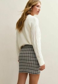 Woman with long blonde hair wearing a white cable-knit sweater and a black-and-white checkered mini skirt, facing sideways.