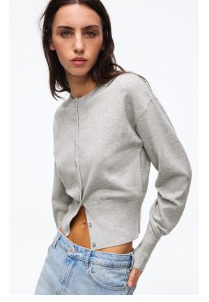 Jonge vrouw die een lichtgrijs cropped vest met knopen en lichtblauwe jeans met hoge taille draagt, poseert tegen een witte achtergrond.