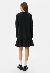 Musta pitkähihaine mekko, jossa on röyhelöhelma, avainaukko selässä ja sileä pinta, yhdistettynä mustiin platform-kengiin ja valkoisiin sukkiin.