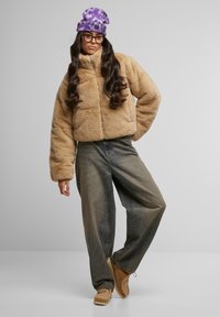 Beige faux pelsjakke med høy krage, kombinert med løse, mørk denimjeans og brune semskede sko. Lilla tie-dye lue og briller fullfører stilen.