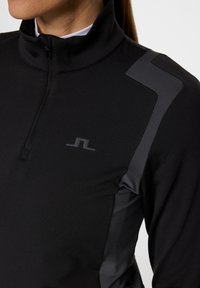 Schwarze Sportjacke mit Reißverschluss und hohem Kragen, die graue Akzente an den Schultern hat und ein schlankes, tailliertes Design aufweist. Weiche, glatte Textur.