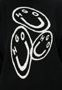 Zwarte trui met grote witte cirkelvormige logo's met de letters "HGO" en glimlachende gezichten. Gebreide textuur met een geribde halslijn en zoom.