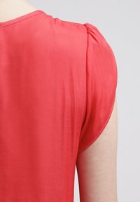 Nahaufnahme einer Person, die eine rote Bluse mit kurzen Ärmeln trägt und den oberen Rücken sowie die Schulter zeigt.