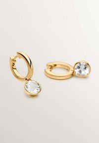 Pendientes de aro en tono dorado con un colgante de cristal ovalado y transparente suspendido de cada aro. Acabado liso con una textura pulida.