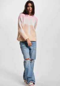 Lys rosa og beige genser med dryppmønster, lange ermer og ribbestrikket halslinning, sammen med lyseblå slitte jeans og casual sko.