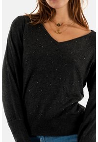 Schwarzer V-Ausschnitt-Pullover mit langen Ärmeln, der kleine silberne Verzierungen und gerippte Bündchen am Saum aufweist. Weiche Strickstruktur.