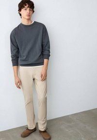 Grauer Pullover, beige Hose, braune Schuhe. Glatte Textur, lässiges Design, taillierte Silhouette, minimalistischer Stil. Stehend vor hellem Hintergrund.