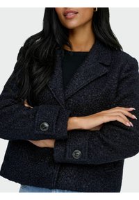 Manteau noir texturé avec un design à col, des poignets boutonnés et une coupe carrée. Gros boutons visibles et texture de tissu bouclée douce.