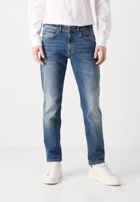 Blue denim jeans with a straight cut, featuring a five-pocket design, contrast stitching, and a faded finish. Paired with white sneakers.