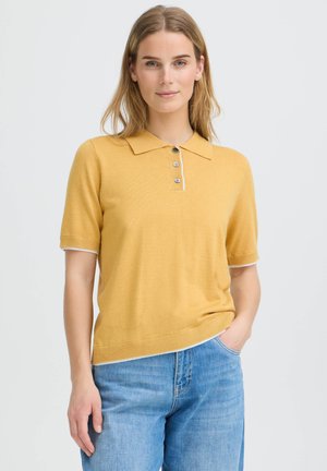 Femme aux cheveux blonds portant un polo jaune moutarde à manches courtes et un jean bleu, debout avec une main dans la poche sur un fond uni.