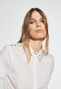 Camicia bianca con bottoni e borchie dorate sulle spalle e sul colletto. Tessuto fresco, design classico e vestibilità strutturata.