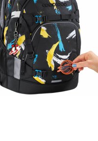 Schwarzer Rucksack mit abstrakten blauen, gelben und weißen Mustern. Enthält einen kleinen charmanten roten und weißen Turnschuh sowie ein orangefarbenes rundes Etikett.