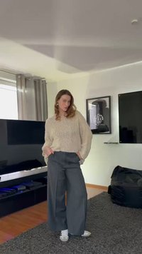 Béžový pletený sveter, široké sivé nohavice a biele tenisky. Neformálny outfit s voľným strihom, zobrazený v svetlom vnútornom prostredí.