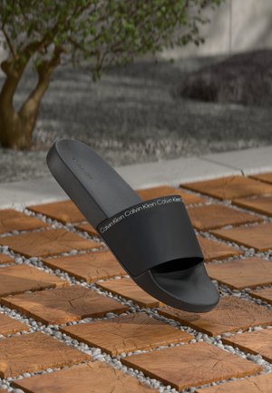 Black rubber slide sandal with a wide strap featuring silver lettering, resting above textured wooden tiles with pebbles between them.