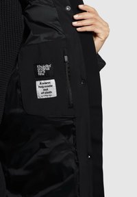 Zwarte outerwear met een gladde, glanzende binnenkant, met etiketten en een ritszak aan de linkerkant. De stof lijkt duurzaam, warm en waterafstotend.
