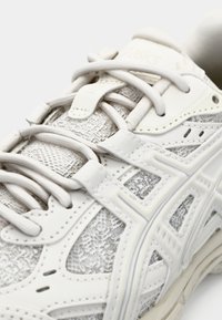 White athletic shoe featuring textured mesh overlay, reinforced heel, and distinct logo accents. Laces in a neutral tone, streamlined design.