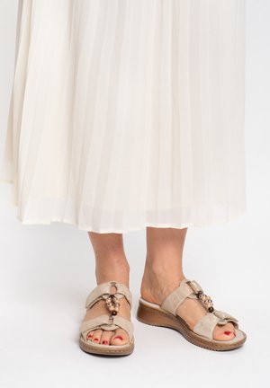 Pies con sandalias cuña beige adornadas con cuentas decorativas, combinadas con una falda plisada blanca, sobre un fondo blanco.