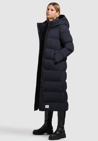 Dlhý námornícky puffer kabát s kapucňou, vybavený gombíkovým zapínaním, zipsovými vreckami a detailom loga na spodnom leme. Hladká, prešívaná textura.