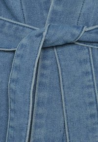 Denim tkanina v svetlo modri barvi z zavezanim pasom. Ponuja vidno šivanje in gladko teksturo. Poudarek na detajlu zavezovanja pasu.