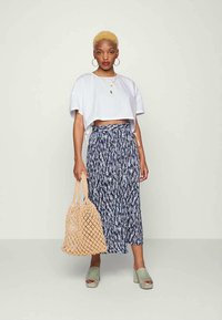 Mulher com cabelo loiro curto, vestindo um top branco cropped, saia midi padrão azul-marinho, sandálias plataformas verdes, segurando uma grande bolsa de palha tecida.