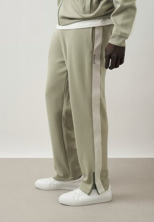 Uomo che indossa pantaloni della tuta beige con strisce laterali e cerniere alle caviglie, abbinati a sneakers bianche e top coordinato.