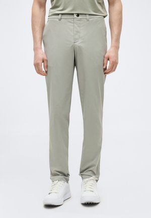 Čovjek koji nosi svijetlosive ravne hlače i bijele tenisice s vezicama, stoji nasuprot jednostavnoj svijetloj pozadini.