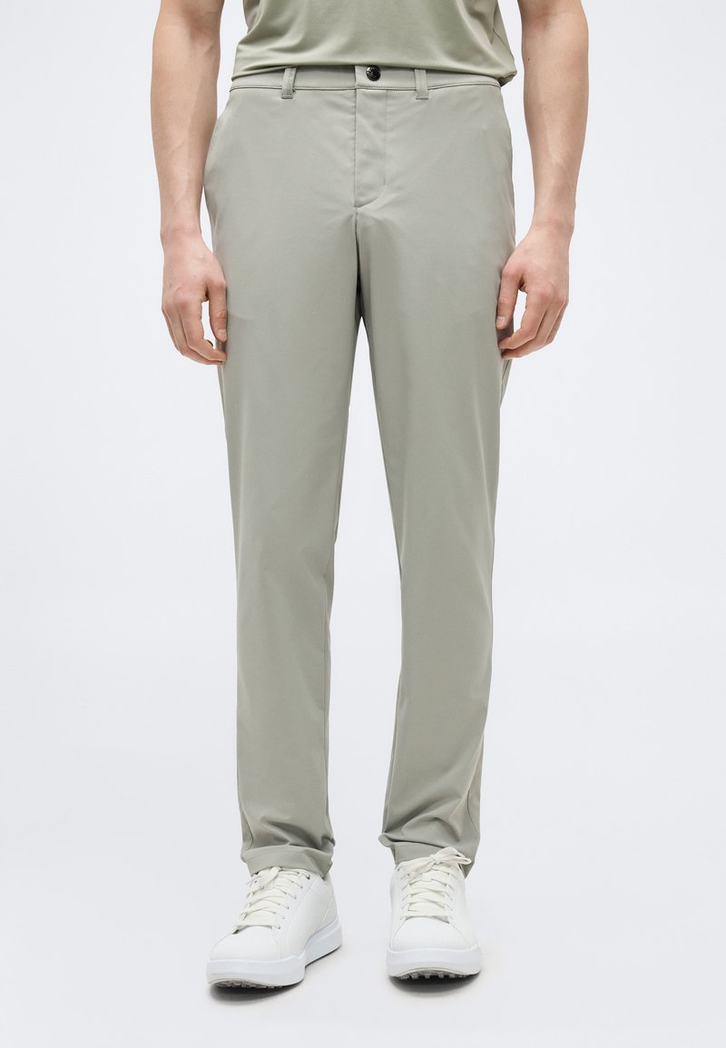 Homme portant un pantalon gris clair à jambes droites et des baskets blanches à lacets, debout devant un fond uni clair.