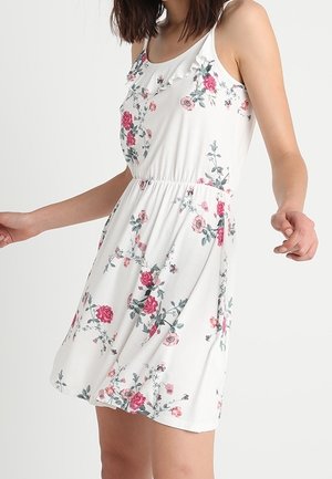 Vrouw draagt een mouwloze witte jurk met roze en groene bloemenprint, elastische taille en een gedetailleerde ruches aan de halslijn.