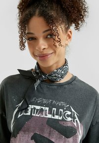 Zwarte graphic t-shirt met een vervaagde print van Metallica, gecombineerd met een zwarte sjaal met witte patronen. Krullend haar omlijst het gezicht.