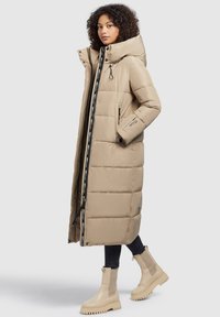 Geacă puffer bej cu guler înalt, fermoar frontal și bandă de branding în contrast. Două buzunare laterale și un design până la genunchi. Textură netedă.