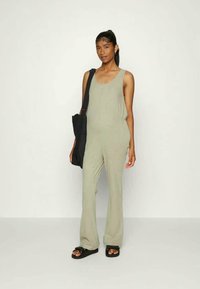 Macacão cáqui com pernas largas, design sem mangas, textura suave e um corte descontraído, combinado com uma mala preta de ombro e sandálias deslizantes.