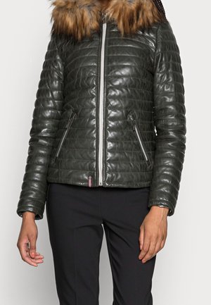Femme portant une veste matelassée vert foncé avec un col en fourrure et des poches zippées, associée à un pantalon noir.