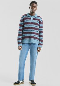 Sweter w paski w odcieniach fioletu i niebieskiego, wykonany z puszystego materiału, z kołnierzykiem, trzema guzikami i ściągaczami na rękawach. W połączeniu z jasnymi dżinsami.